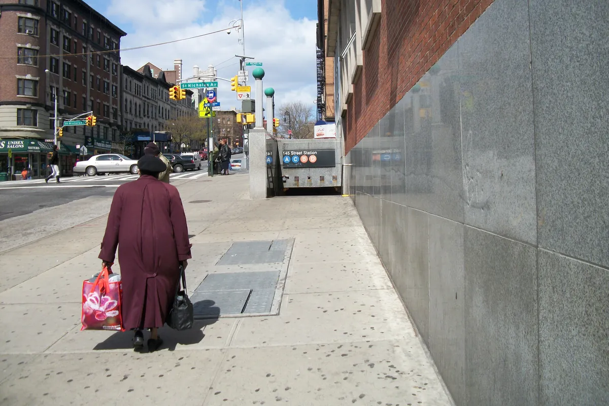 Un ingresso della metropolitana all'angolo di una strada di Harlem, con una donna che porta borse della spesa che passa davanti a alti palazzi di mattoni